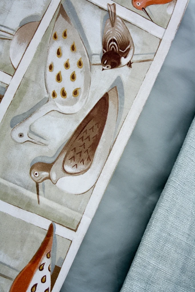 Pale blue fabric printed with illustrated birds and feathers beside a plain blue textile